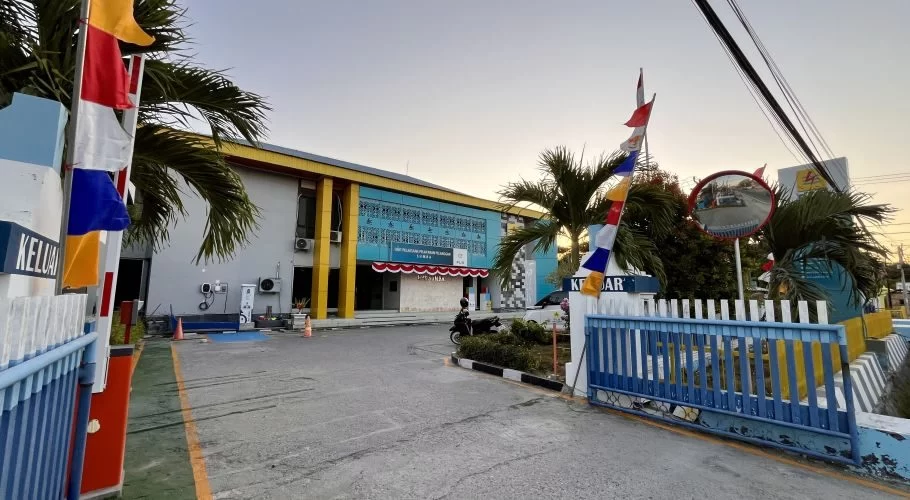 The PLN Office in Waingapu features a blue and yellow building with a driveway, blue and white gates, colorful flags, and palm trees under a clear sky.
