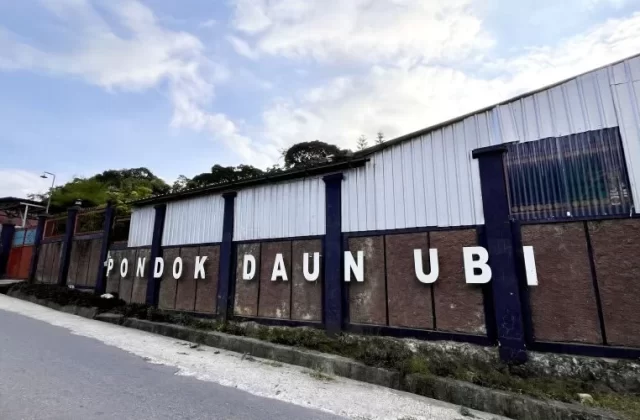 The white letters 'PONDOK DAUN UBI' are displayed on a brown textured wall with dark blue pillars, beneath a white corrugated metal building and a cloudy sky, seen from a paved road.