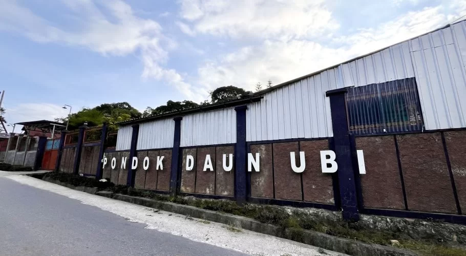 The white letters 'PONDOK DAUN UBI' are displayed on a brown textured wall with dark blue pillars, beneath a white corrugated metal building and a cloudy sky, seen from a paved road.