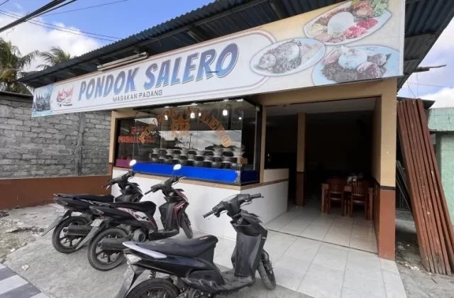 The exterior of Pondok Salero restaurant in Waikabubak features a white sign with blue text, a corrugated roof, and parked motorbikes under a bright sky.