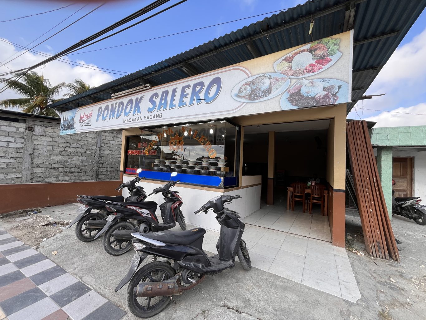 The exterior of Pondok Salero restaurant in Waikabubak features a white sign with blue text, a corrugated roof, and parked motorbikes under a bright sky.