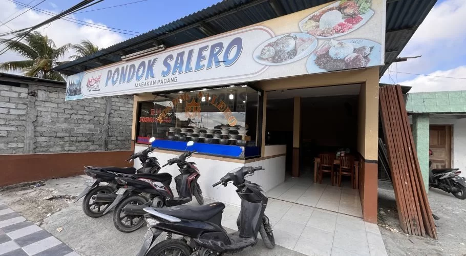 The exterior of Pondok Salero restaurant in Waikabubak features a white sign with blue text, a corrugated roof, and parked motorbikes under a bright sky.