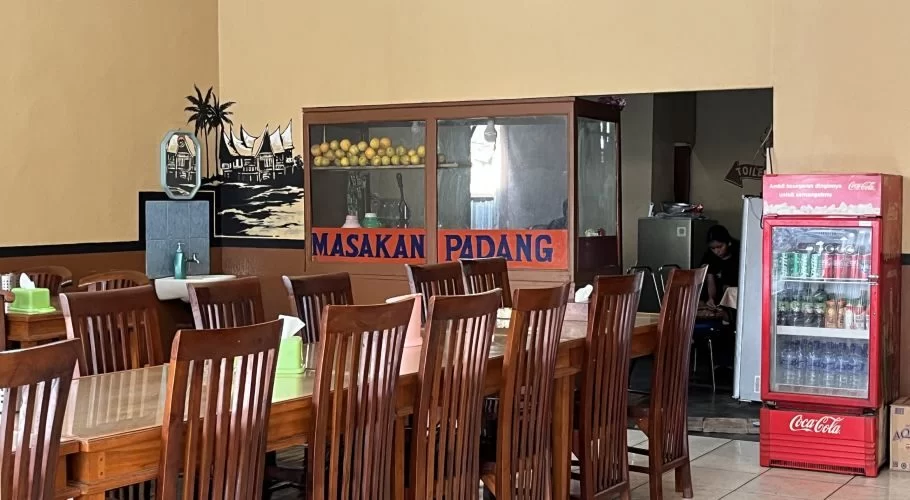 The interior of Pondok Salero restaurant in Waikabubak shows long wooden tables and chairs, a Padang cuisine display, a handwashing station, and a beverage cooler against tan walls.