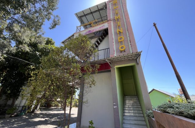 A multi-story reddish-pink and grey building, Pondok Wisata & Cafe Elim, stands under a clear blue sky, surrounded by green trees in East Sumba.