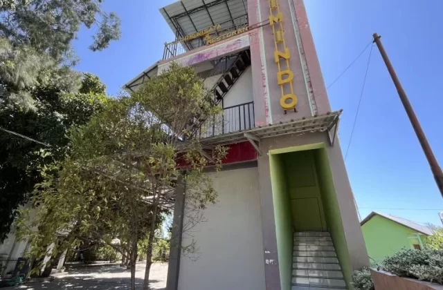 A multi-story reddish-pink and grey building, Pondok Wisata & Cafe Elim, stands under a clear blue sky, surrounded by green trees in East Sumba.