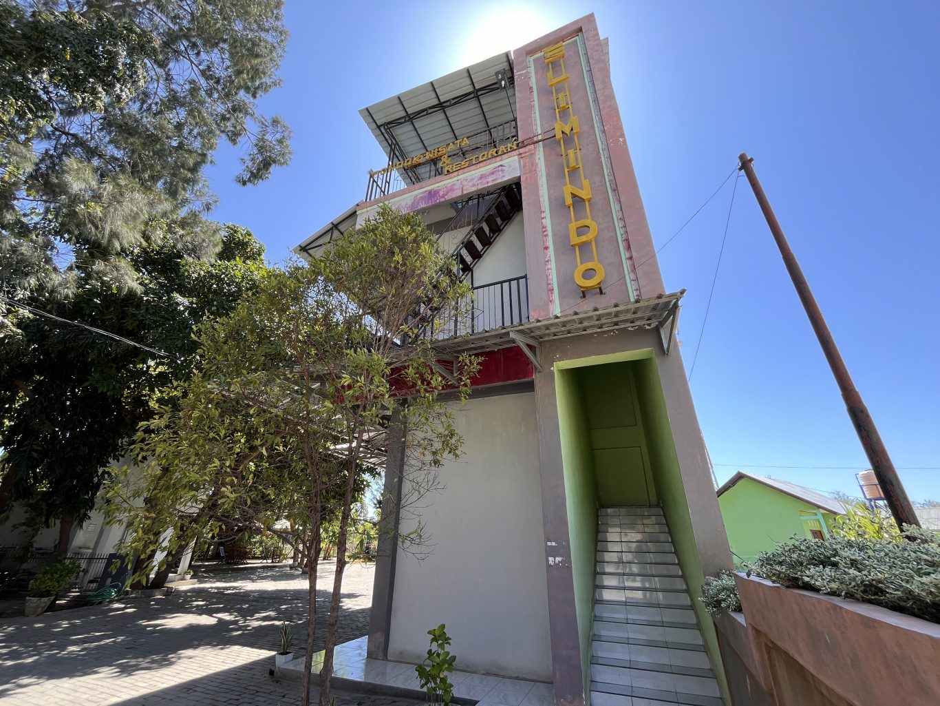 A multi-story reddish-pink and grey building, Pondok Wisata & Cafe Elim, stands under a clear blue sky, surrounded by green trees in East Sumba.