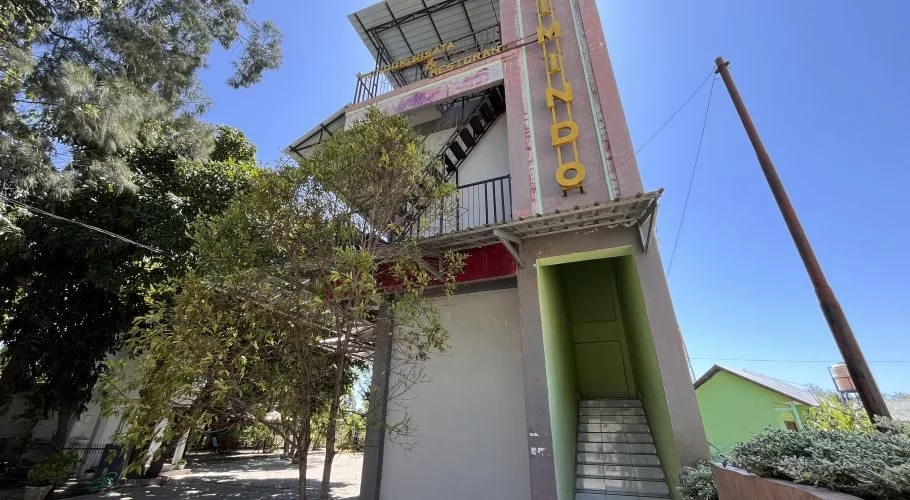 A multi-story reddish-pink and grey building, Pondok Wisata & Cafe Elim, stands under a clear blue sky, surrounded by green trees in East Sumba.