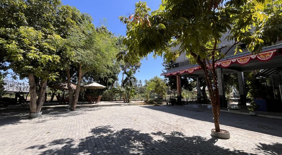 A spacious paved outdoor area at Pondok Wisata & Cafe Elim is shaded by numerous green trees under a clear blue sky, with a white umbrella and seating visible.