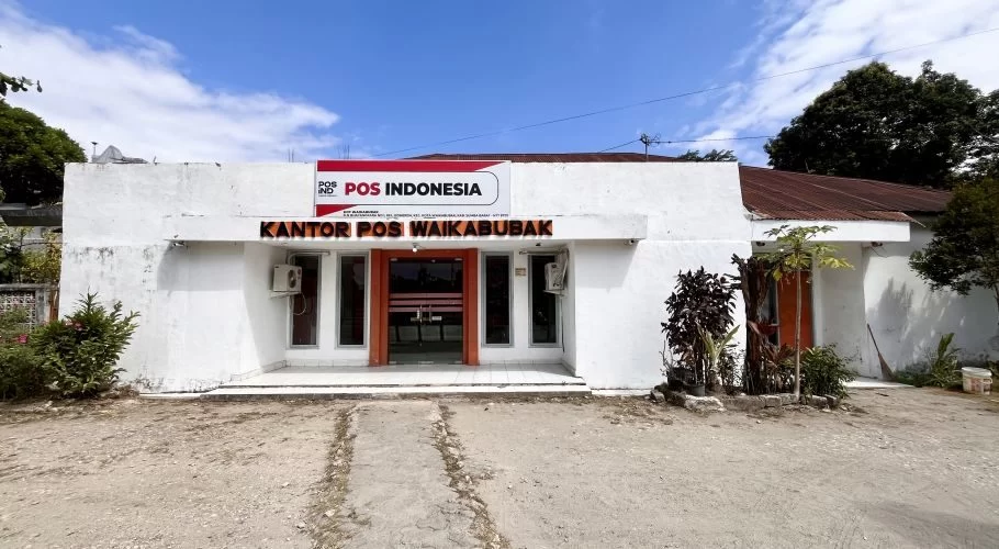 The white Pos Indonesia post office building in Waikabubak, Sumba, features a brown entrance and windows, with small plants in front under a partly cloudy blue sky.