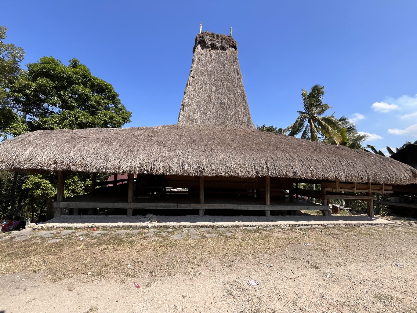 A traditional Sumba house with a distinctive tall, conical thatched roof and a wide lower thatched section stands under a clear blue sky.