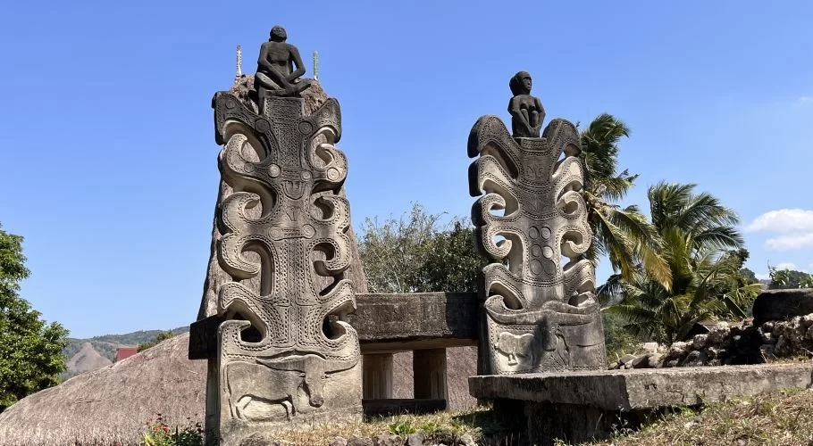 Two large, grey, intricately carved stone statues with seated human figures on top stand under a bright blue sky in Praibakul Village, Sumba.