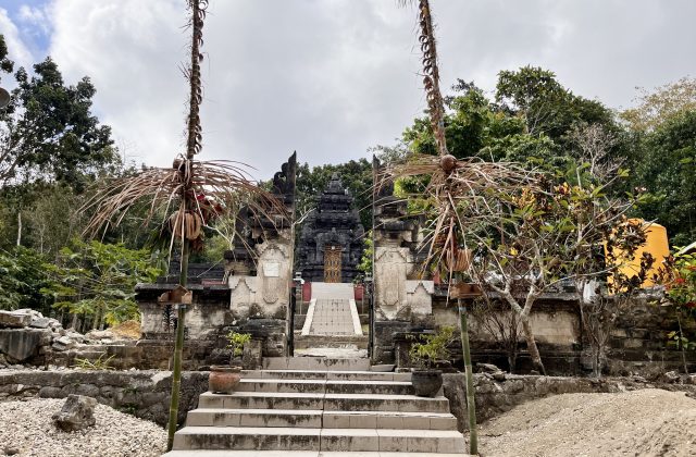 A dark stone Hindu temple entrance with steps and a multi-tiered structure is framed by decorated poles and green trees.