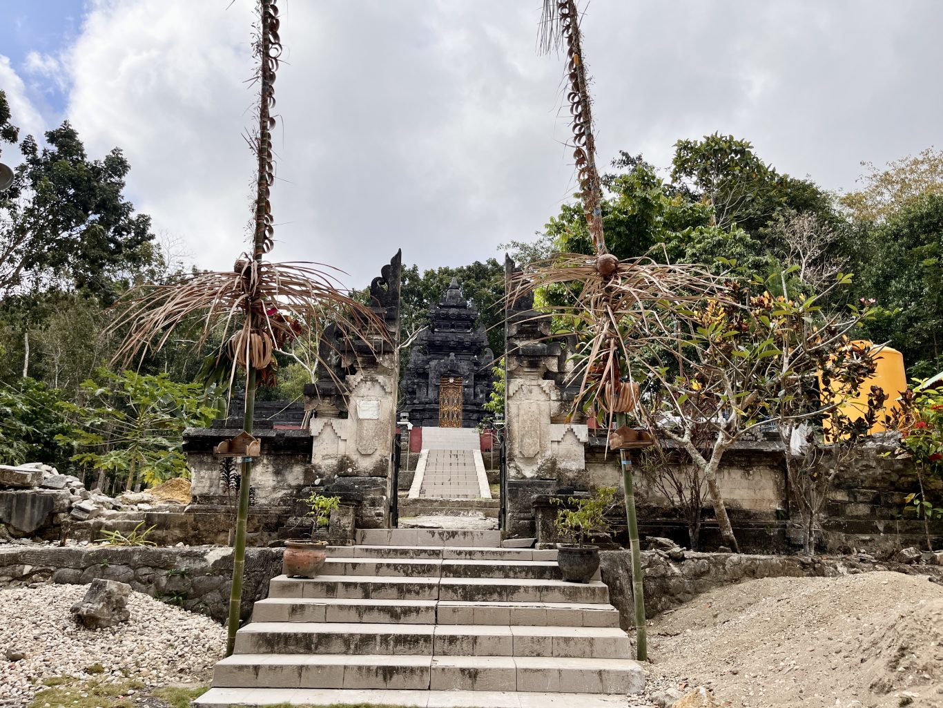 A dark stone Hindu temple entrance with steps and a multi-tiered structure is framed by decorated poles and green trees.