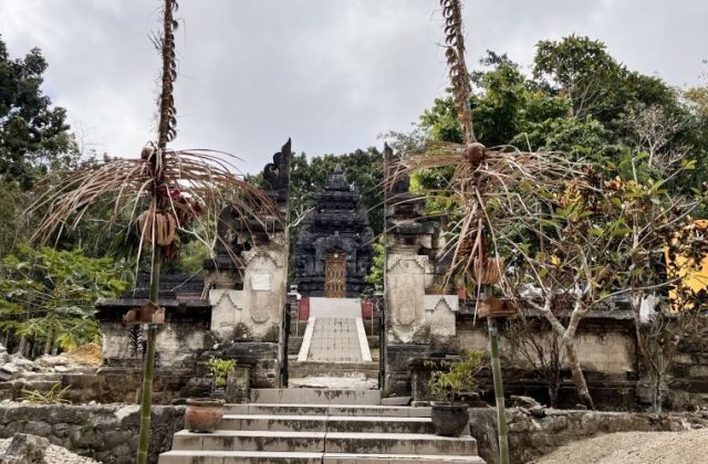 A dark stone Hindu temple entrance with steps and a multi-tiered structure is framed by decorated poles and green trees.