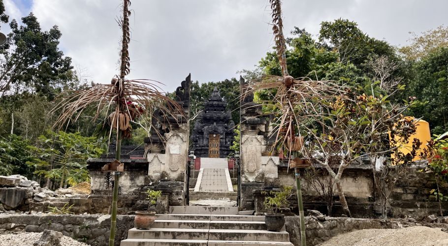 A dark stone Hindu temple entrance with steps and a multi-tiered structure is framed by decorated poles and green trees.