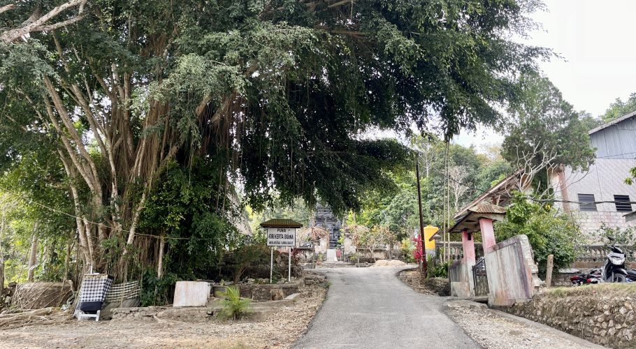 A paved road leads to the entrance of Pura Giri Kertha Bhuana, flanked by a massive banyan tree on the left and buildings on the right, under a bright sky.