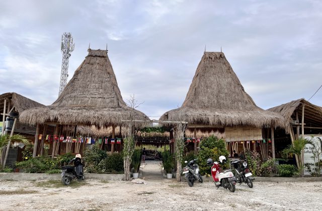 Two large traditional Sumba houses with tall, conical thatched roofs stand prominently, flanked by smaller thatched structures and lush green plants. Several motorbikes are parked on a dirt lot in front, under a cloudy sky, with a cell tower visible on the left.