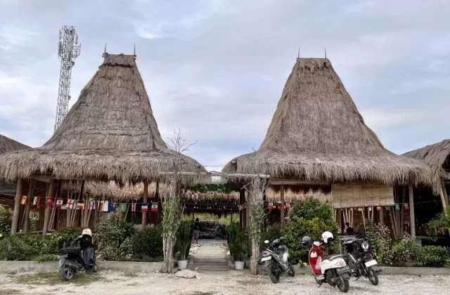 Two large traditional Sumba houses with tall, conical thatched roofs stand prominently, flanked by smaller thatched structures and lush green plants. Several motorbikes are parked on a dirt lot in front, under a cloudy sky, with a cell tower visible on the left.