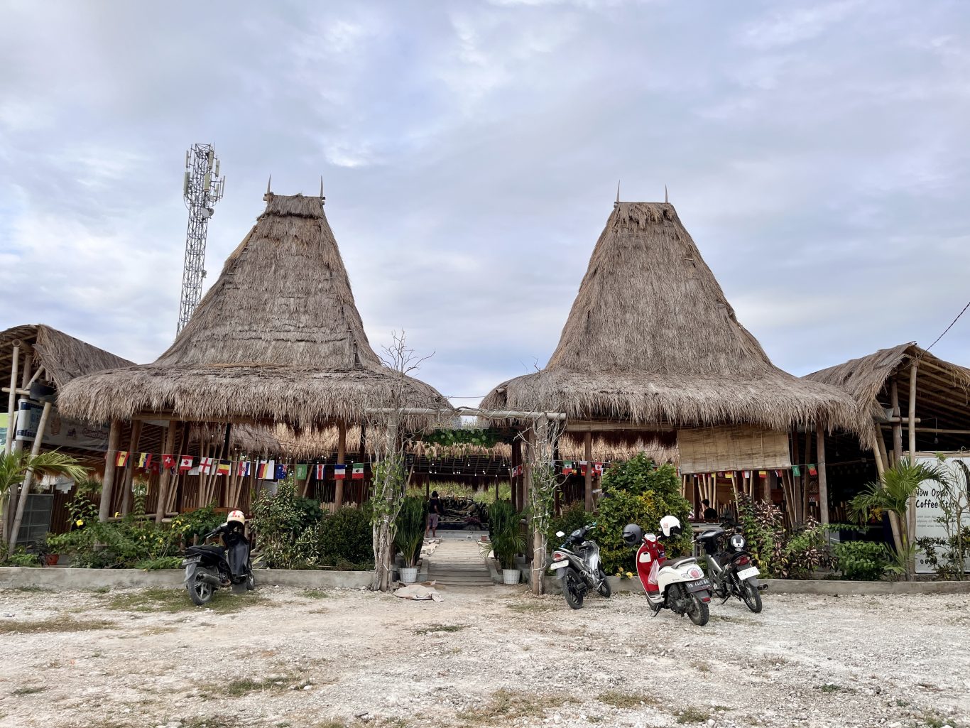 Two large traditional Sumba houses with tall, conical thatched roofs stand prominently, flanked by smaller thatched structures and lush green plants. Several motorbikes are parked on a dirt lot in front, under a cloudy sky, with a cell tower visible on the left.