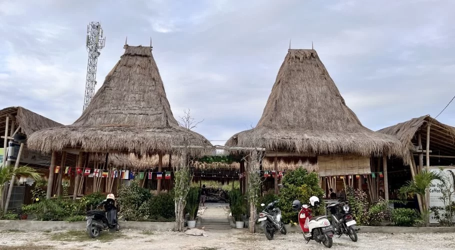 Two large traditional Sumba houses with tall, conical thatched roofs stand prominently, flanked by smaller thatched structures and lush green plants. Several motorbikes are parked on a dirt lot in front, under a cloudy sky, with a cell tower visible on the left.