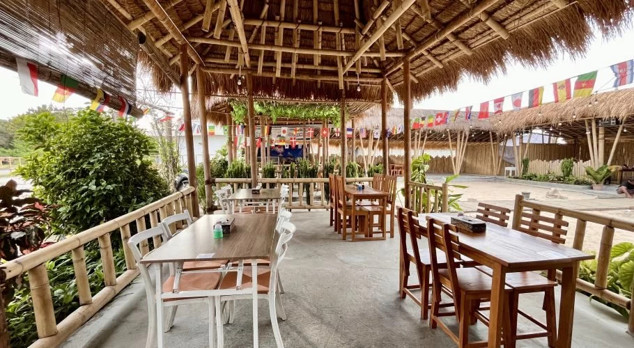 An open-air cafe features a thatched bamboo roof, wooden tables, and chairs, with colorful international flags strung overhead and green foliage around the perimeter.