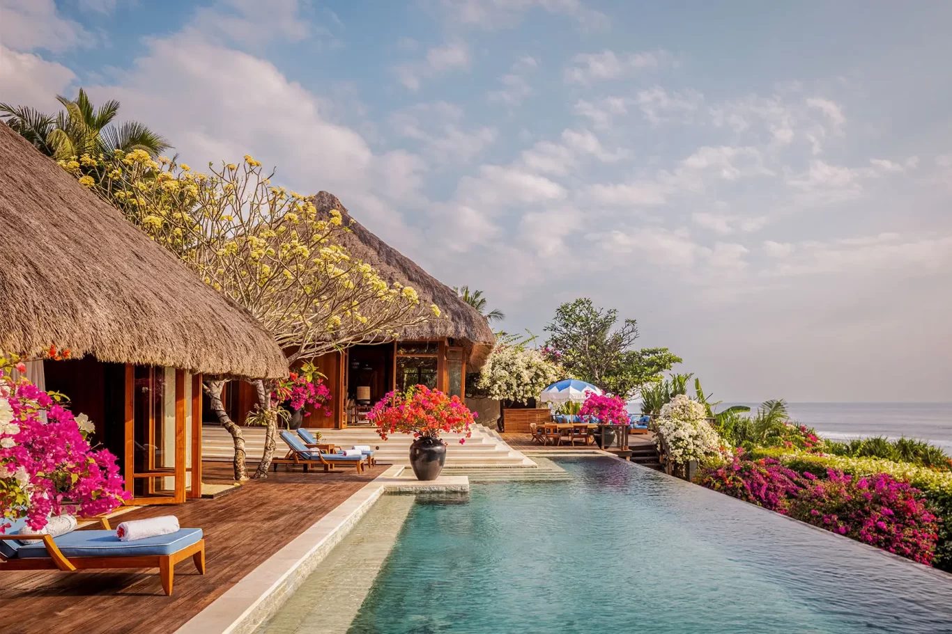 Thatched-roof bungalows, an infinity pool, and vibrant bougainvillea overlook the blue ocean under a partly cloudy sky.