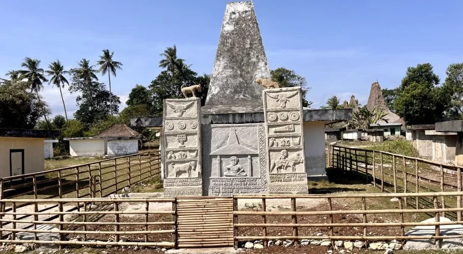 Visible is a large, grey stone ancestral grave monument with detailed carvings and two animal statues on top, enclosed by a bamboo fence, set in a traditional village with palm trees under a blue sky.