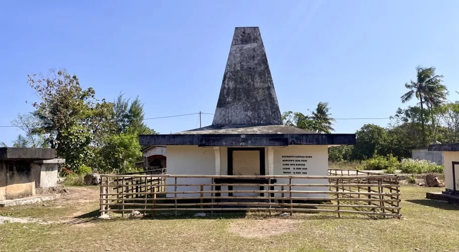 A white building with a tall, dark, pyramid-shaped roof stands in a grassy field, surrounded by a bamboo fence and other stone graves under a clear blue sky.