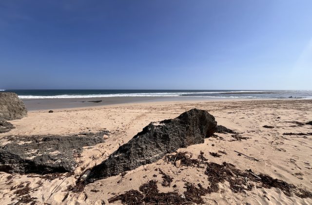 Ratenggaro Beach features light brown sand, dark rocky formations in the foreground, and clear blue ocean waves under a bright blue sky.