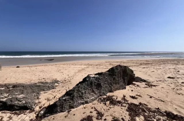 Ratenggaro Beach features light brown sand, dark rocky formations in the foreground, and clear blue ocean waves under a bright blue sky.
