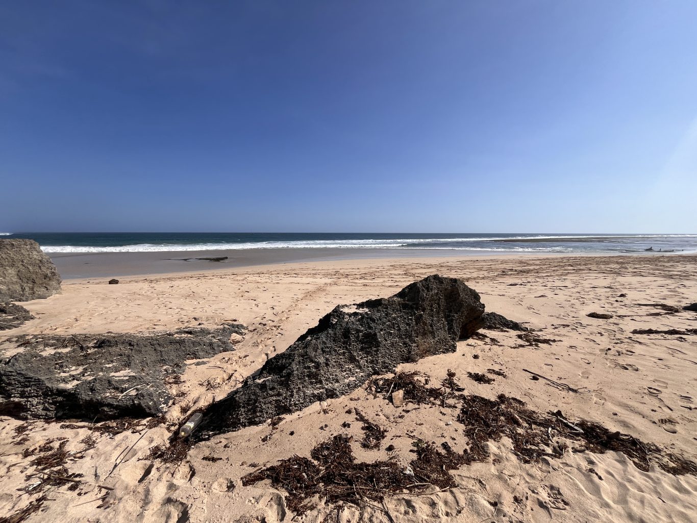Ratenggaro Beach features light brown sand, dark rocky formations in the foreground, and clear blue ocean waves under a bright blue sky.