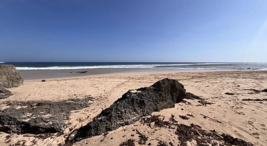 Ratenggaro Beach features light brown sand, dark rocky formations in the foreground, and clear blue ocean waves under a bright blue sky.