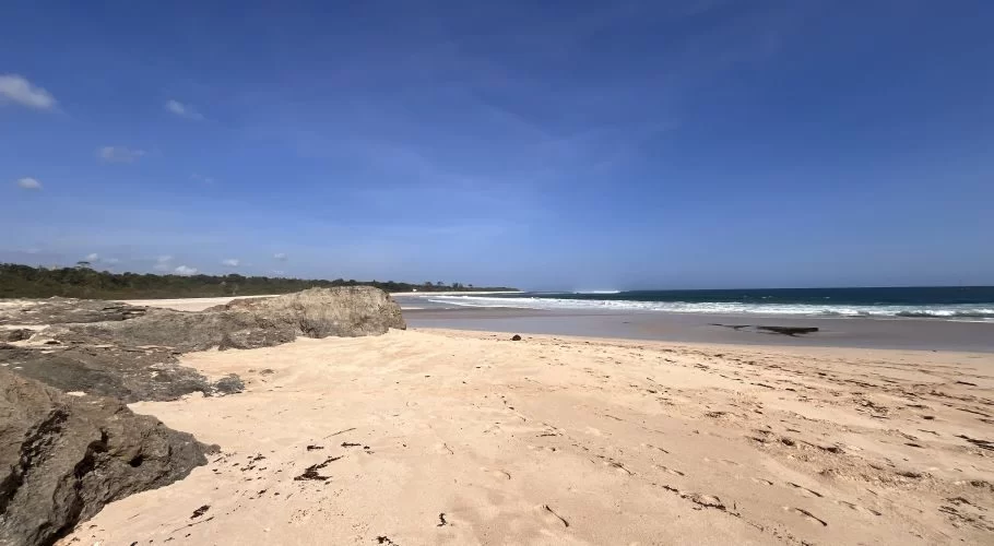A wide view of Ratenggaro Beach shows light brown sand, dark rocky formations, and clear blue ocean waves under a bright blue sky.