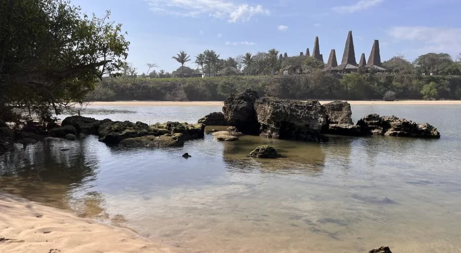 A serene view of Ratenggaro Beach with light brown sand, clear shallow water, dark rocks, and traditional Sumba houses with tall roofs on a distant green bank under a blue sky.