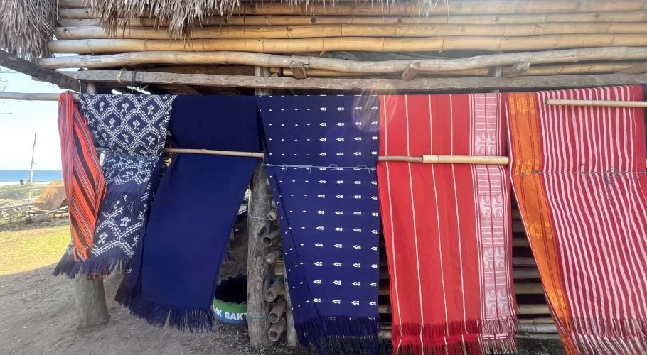 Colorful traditional Sumba ikat textiles hang from bamboo poles under a thatched roof, with the ocean visible in the background.