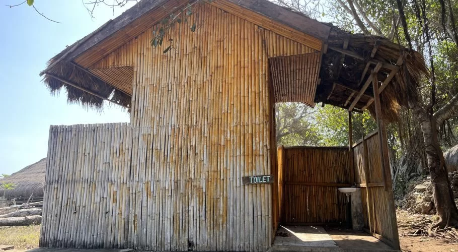 A rustic outdoor toilet building with bamboo walls and a thatched roof, featuring a "TOILET" sign, stands under a clear sky near trees.