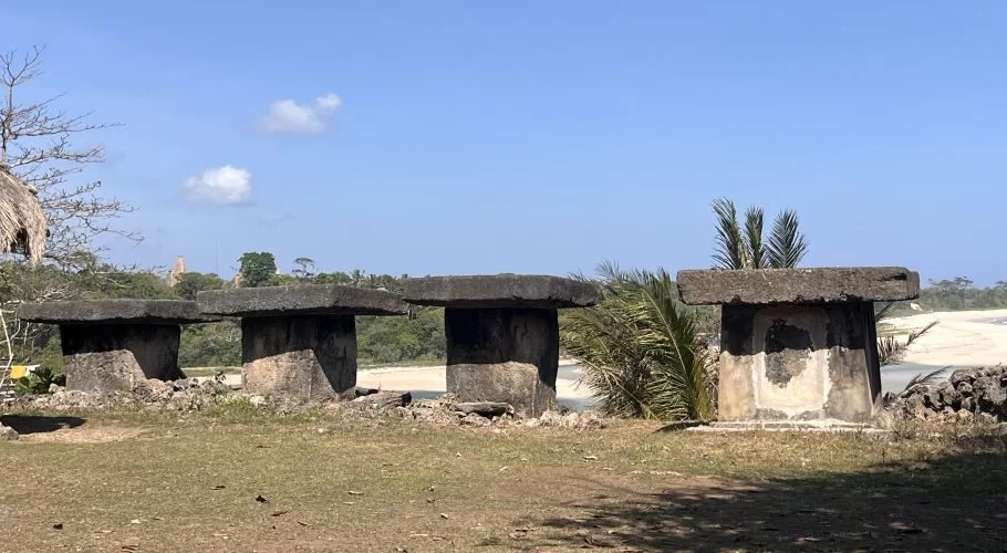 Four ancient, dark grey megalithic stone tombs stand on dry grass, overlooking a sandy riverbank and green trees under a clear blue sky.