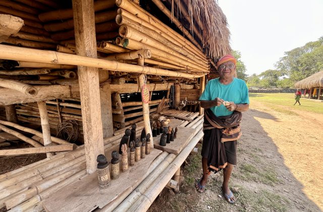 An elderly Sumba artisan in a turquoise shirt stands outside a traditional bamboo home, holding a carving, with wooden statues displayed.