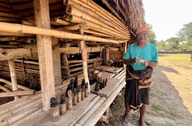 An elderly Sumba artisan in a turquoise shirt stands outside a traditional bamboo home, holding a carving, with wooden statues displayed.