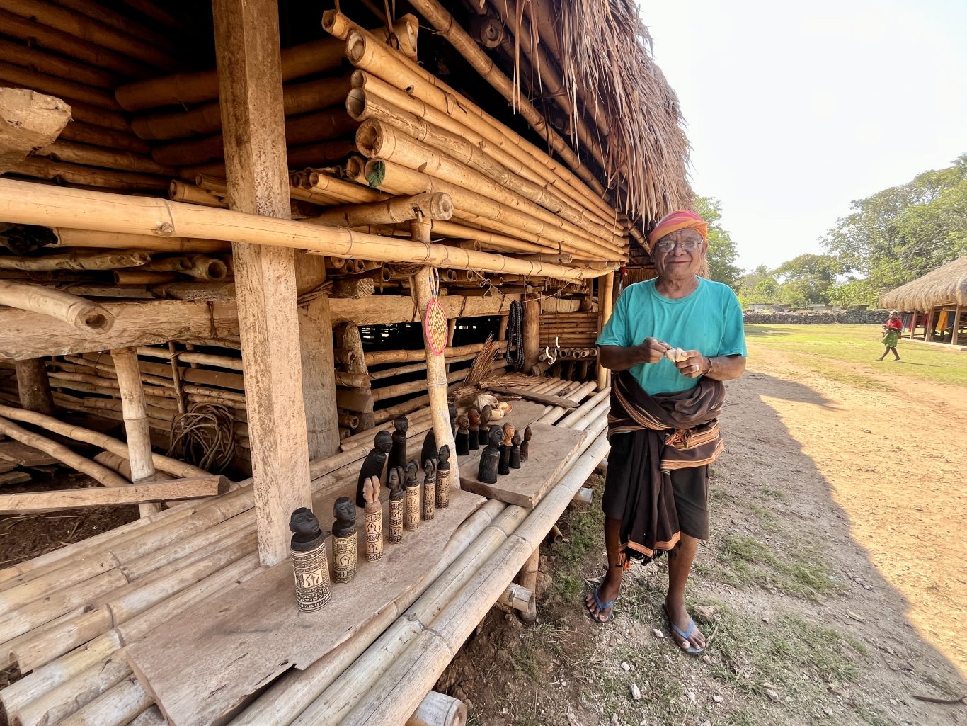 An elderly Sumba artisan in a turquoise shirt stands outside a traditional bamboo home, holding a carving, with wooden statues displayed.