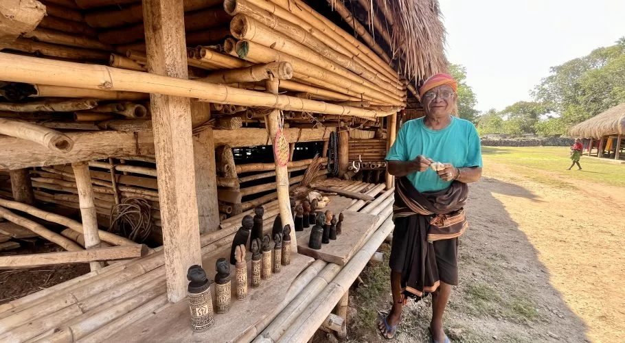 An elderly Sumba artisan in a turquoise shirt stands outside a traditional bamboo home, holding a carving, with wooden statues displayed.