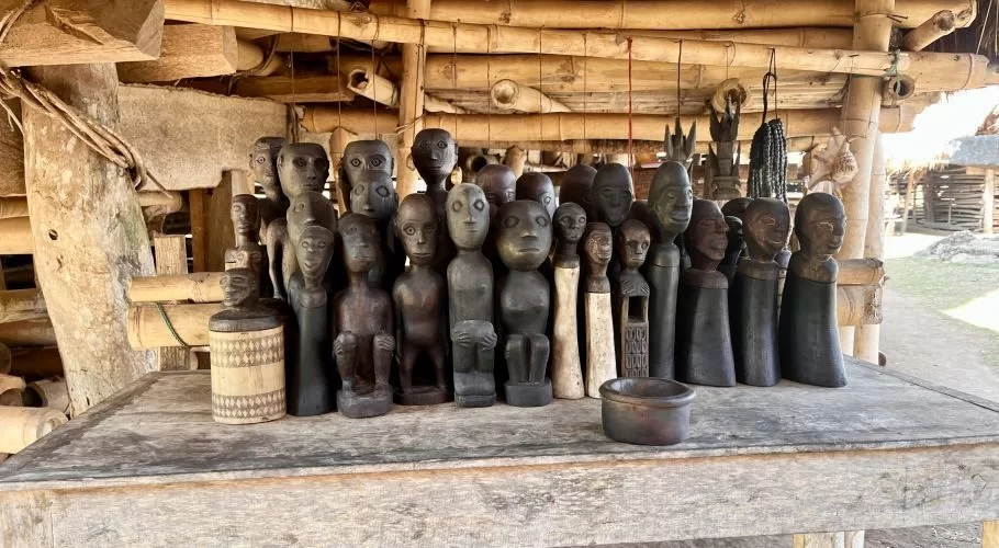A collection of dark and light wooden human-like sculptures and a patterned container are displayed on a rustic table under a bamboo roof.