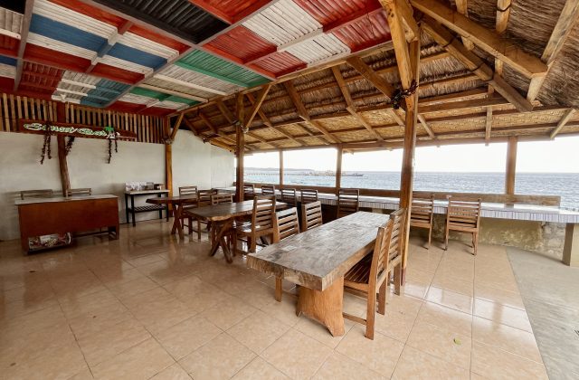 An open-air restaurant with wooden tables and chairs, a tiled floor, and a ceiling of colorful panels and thatch, overlooks the blue ocean.