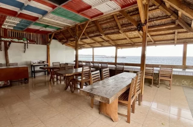 An open-air restaurant with wooden tables and chairs, a tiled floor, and a ceiling of colorful panels and thatch, overlooks the blue ocean.