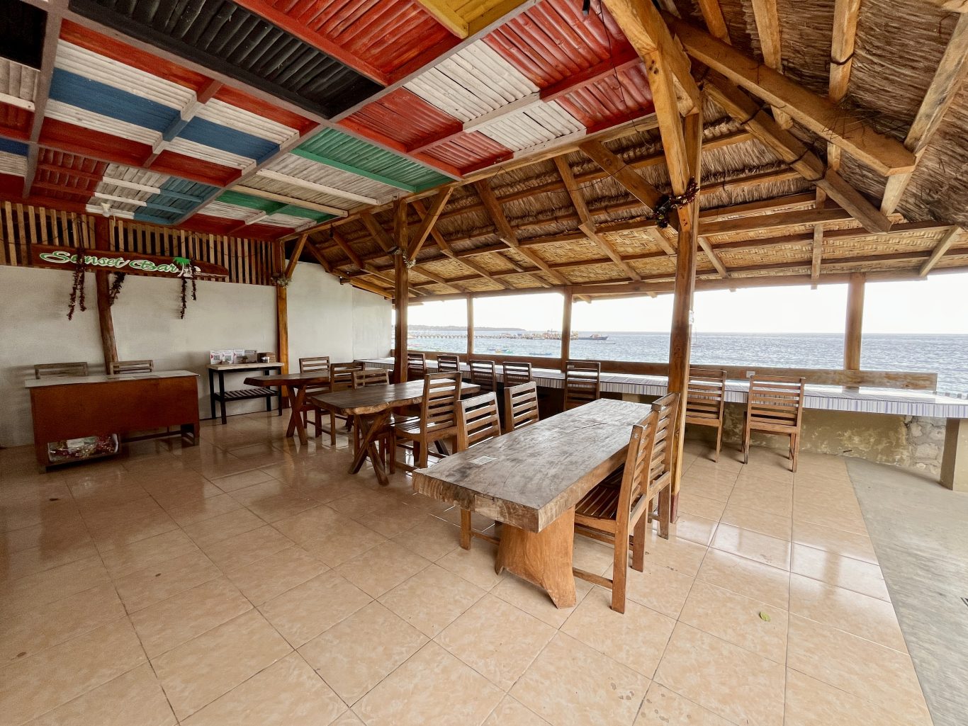 An open-air restaurant with wooden tables and chairs, a tiled floor, and a ceiling of colorful panels and thatch, overlooks the blue ocean.