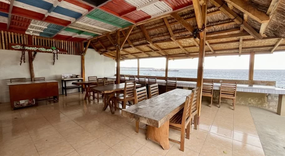 An open-air restaurant with wooden tables and chairs, a tiled floor, and a ceiling of colorful panels and thatch, overlooks the blue ocean.
