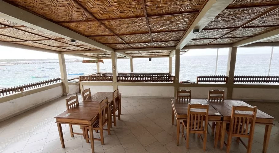 Wooden tables and chairs fill an open-air restaurant with a woven ceiling, offering views of the blue ocean dotted with boats.