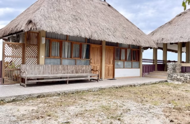 A traditional Redemptorist Villa building with a large thatched roof and wooden features sits by the ocean under a cloudy sky, with a long wooden bench on its porch.