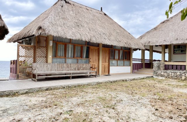 A traditional Redemptorist Villa building with a large thatched roof and wooden features sits by the ocean under a cloudy sky, with a long wooden bench on its porch.