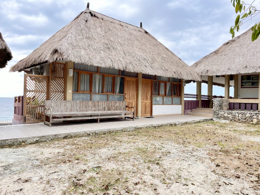 A traditional Redemptorist Villa building with a large thatched roof and wooden features sits by the ocean under a cloudy sky, with a long wooden bench on its porch.